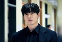 Homem de cabelo preto e roupa escura em um corredor, com pessoas desfocadas ao fundo e preenchimento lateral desfocado.