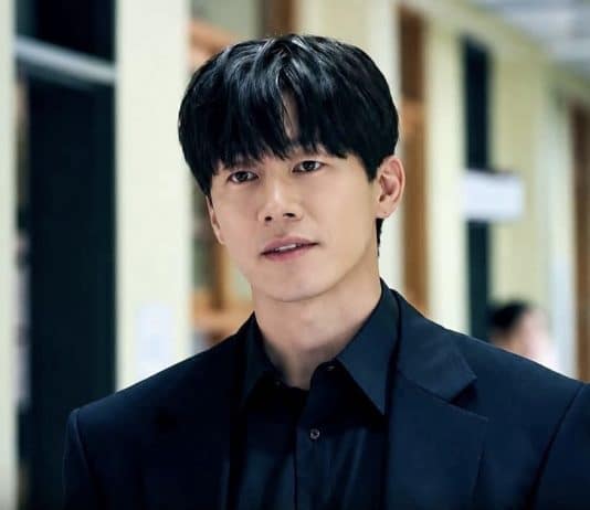 Homem de cabelo preto e roupa escura em um corredor, com pessoas desfocadas ao fundo e preenchimento lateral desfocado.